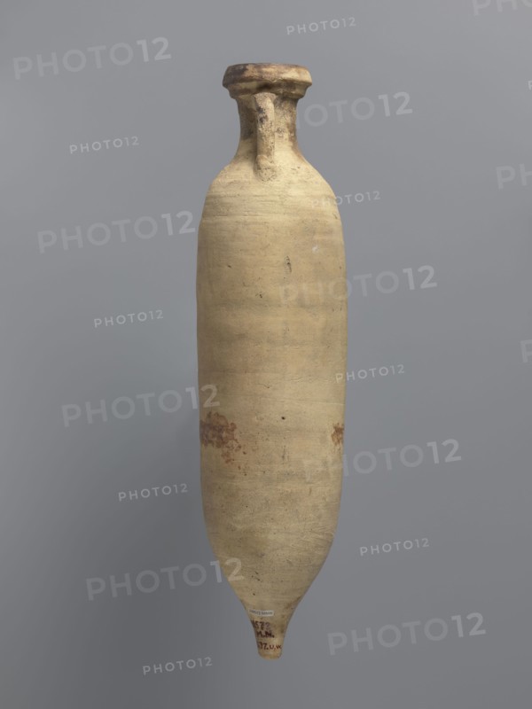 Amphora spatheion, unknown North African workshop Tunisia/Libya. Cylindrical, elongated amphora with a smooth yellow-green surface, probably covered with a coating produced during the firing process natural engobe, self-slip. Two-stage profiled spout with a rounded top and an outward-sloping lip. Small loop-shaped handles attached to the shoulders and to the neck just below the spout; conical bottom. Inner walls covered with a resin impregnation, also visible on the outer walls of the upper part of the amphora. Complete vessel; ceramic body not visible. Date: 2nd half of the 6th–7th century. Provenance: Edfu. Function/type: vessel, amphora spatheion. Dimensions: height 44.2 cm; rim diameter 6.6 cm; body diameter 10 cm., A tall, narrow, elongated pottery vessel with a rounded shoulder, slim neck, and a flared, slightly worn rim, shown against a plain gray background; the vessel surface is predominantly beige with areas of light tan and small patches of darker brown discoloration and spo...
