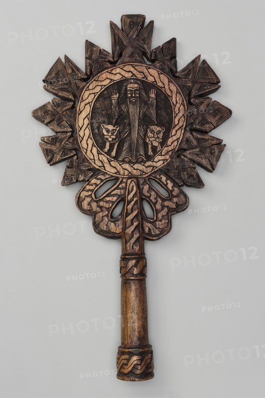 Processional cross, Ethiopian workshop. In the center of the cross, in the tondo, a standing man is depicted frontally with hands raised in an orant gesture. On his sides stand a lion and a lioness. The man has a long, ankle-length beard—likely the hermit Paulos. The tondo is bordered by a wide braid-decorated band from which radiate small crosses with rhomboid arms. The short, sleeve-shaped handle is decorated with two rings with a braided ornament. The reverse shows a winged figure above two birds. 20th century, Ethiopia. Cross; height 44 cm, width 24 cm., A carved wooden object with a long cylindrical handle and an ornate top featuring a circular medallion surrounded by a radiating geometric design; the carving includes a central figure with two animal faces flanking it, all rendered in carved texture and shallow relief. The surface shows visible carved lines and crosshatching, braided motif bands around parts of the handle, and carved openwork shapes beneath the medallion. The colo...
