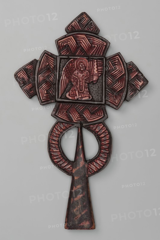 Processional cross, Ethiopian workshop. At the intersection of the cross's arms is a square depicting three seated, frontally posed figures of the Holy Trinity, each holding a spherical object in their right hand as a symbol of power over the world. The arms of the cross are trapezoidal with rounded outer sides; the horizontal beam terminates in trapezoidal crosses, and a similar cross is placed at the top of the vertical beam. The handle is sleeve-shaped, widening at the bottom, with two semicircles attached to its sides. On the reverse, at the intersection of the arms, there is an archangel holding a sword. 20th century, Ethiopia. Material: metal cross. Height 39 cm, width 23 cm., An intricately carved wooden object with a cross-like top section and a long tapered handle, featuring a central square panel containing a carved human figure with wings; the surface shows carved geometric patterns and textured areas, with visible colors including dark brown, reddish-brown, black, and a pal...