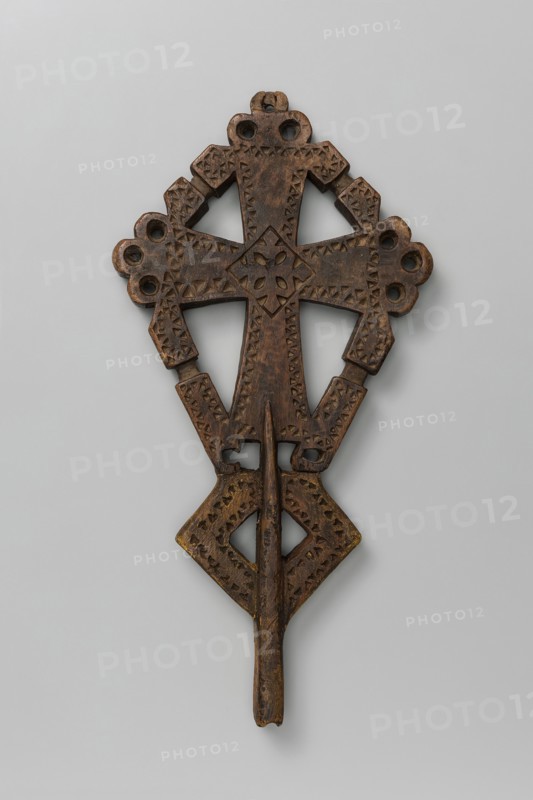 Processional cross, Ethiopian workshop. A small, likely processional isosceles cross with slightly widening arms terminating in trefoil-shaped projections except for the lower arm, which has a mounting socket. Arms are connected by crossbars with a central recess on each. Surface decorated with incised small triangles forming an edge ornament. At the arms' intersection is a rhombus containing a rosette and central cross; lower part features a rhombus through which the socket runs, its edges likewise decorated with triangular incisions. 20th century, Addis Ababa. Height 29 cm, width 14.5 cm., A carved wooden object with a symmetrical, cross-like shape featuring geometric cutouts and incised triangular patterns, displaying a rich brown color with areas of darker brown and hints of yellowish tones.