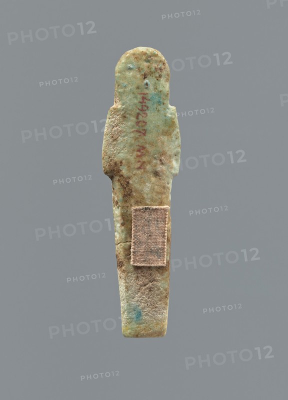 Ushabti, Mummy-shaped figurine, funerary gift, funerary hoe. Hands folded on the chest, uncrossed, right hand under left; both hands hold hoes. Smooth tripartite wig with a headband; facial details poorly legible except for large eyes. Smooth back. No inscription. c. 1069–c. 656 BC Third Intermediate Period, Egypt. Height 6.4 cm., A worn, elongated figurine-like object stands vertically against a plain gray background, showing a rounded head, narrow shoulders, and a tapering body; the surface is rough and pitted with an aged texture, and a small rectangular fabric patch is affixed to the lower torso area. The visible colors include muted green, pale blue, tan, brown, and hints of reddish discoloration, with darker brown spots and lighter sandy areas across the form.