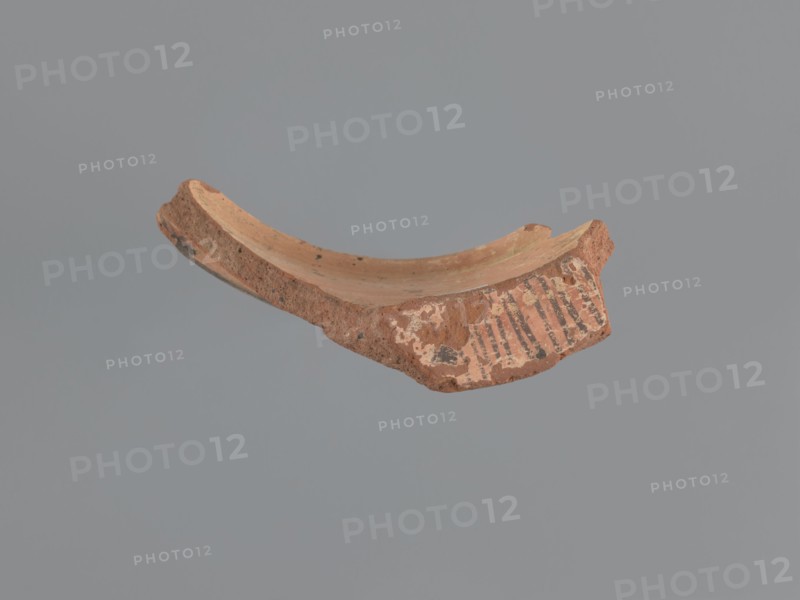 Fragment of a vessel stand, painted Coptic vessel stand with floral decorations. Walls formed into a hoop with the diameter of the upper edge slightly larger than the lower. Decoration painted in black and red on a beige whitewash background. Lower edge bulges outward and inward in the form of a half-cylinder; its outer surface decorated with a black horizontal band. Upper edge has an outward rim; its horizontal outer surface decorated with a red band bearing several black dots and a series of lines perpendicular to the rim. Exterior decorated with almond-shaped, stylized, unevenly sized plant elements with pointed tips upward; each almond-shaped motif filled with a grid of black lines and surrounded by two or three groups of arcuate lines repeating its shape. Made of medium-porous Nile clay. 6th–7th century, Tell Atrib; vessel. Height 8.5 cm, width 13.7 cm, lower outer rim diameter 24 cm, upper outer rim diameter 25 cm., A broken curved fragment of reddish-brown clay with a rough text...