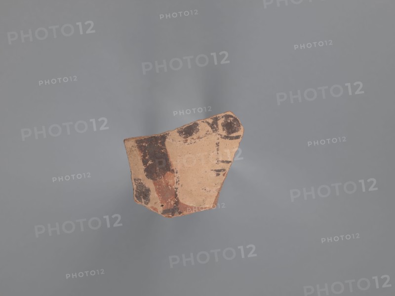 Fragment of the body of a vessel with decoration painted in black and red against a pink-beige engobe background. Around the face, preserved only on the right side of the head, is wavy black hair covering the ear. The black pupils are surrounded by an almond-shaped outline resembling the shape of the eye. The nose is represented by a long vertical and a short horizontal line; the mouth by two unequal lines. A round black dot is next to the face. Nile clay of medium porosity. 5th–6th century, Egyptian workshop, Tell Atrib, painted Coptic vessel with figural decorations; height 3.2 cm, width 4 cm., A fragment of pottery with a rough, irregular shape sits against a plain gray background. The pottery piece is mainly a tan or light brown color with areas of darker brown and reddish-brown pigment forming irregular patches and worn shapes. The surface shows texture variations and some chipped edges, and the lighting creates soft shadows around the fragment.