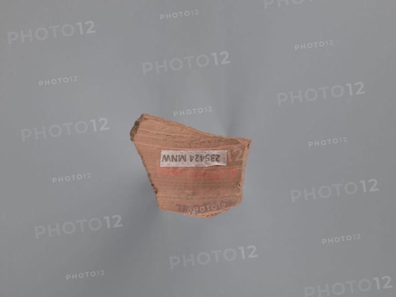 Fragment of a vessel, painted Coptic vessel with figural decoration, Fragment of the body of a vessel with decoration painted in black and red against a pink-beige engobe background. Around the face, preserved only on the right side of the head, is wavy black hair covering the ear. The black pupils are surrounded by an almond-shaped outline resembling the shape of the eye. The nose is represented by a long vertical and a short horizontal line; the mouth by two unequal lines. A round black dot is next to the face. Nile clay of medium porosity. 5th–6th century, Tell Atrib, vessel, height 3.2 cm, width 4 cm, A fragment of reddish-brown pottery with rough, uneven edges sits against a smooth gray background; the pottery piece shows layered texture and slight ridges on its surface and has areas of darker brown along one edge, while small patches of lighter tan appear where the surface is worn.