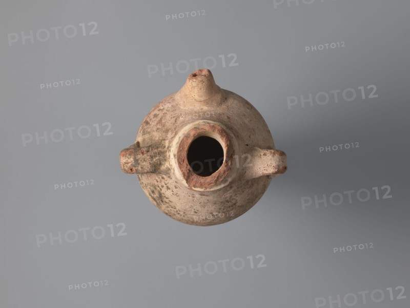 Water vessel qulla, bottles, Coptic vessels, water vessels qullae, qulla, Water vessel bottle, gulla. Ovoid, elongated body, wider in the uppermost part, above half the vessel's height. Low foot, hollow inside, poorly defined externally. Two small vises are glued to the arms, and between them, on an axis perpendicular to their attachment, a conical spout for drinking water is glued. The neck, preserved in its lowest part, is separated from the body by a small, sharply profiled hump. The entire surface of the vessel is covered with light whitewash. Nile clay, medium-porous, 5th century, Tell Atrib, vessel, height 17.0 cm, width 11.5 cm, diameter 4 cm base, A worn round clay vessel viewed from above with a central circular opening and a short tapered spout, two protruding loop handles on opposite sides, a rough textured surface showing patches of darker and lighter areas, and visible surface irregularities; colors include beige, tan, light brown, reddish-brown, and some darker grayish sp...