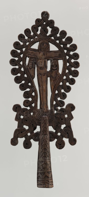 Processional cross, Ethiopian workshop. The pear-shaped frame depicts Christ on the cross with John and Mary standing at the foot in flat relief. Figures are schematically rendered; Christ is bearded, long-haired, with open eyes, head slightly tilted to his right left shoulder, dressed only in a loincloth, feet nailed with a single nail. The pear-shaped frame is decorated with a braided rope pattern and bordered along the outer edge with an openwork braided ornament. The cone-shaped sleeve, flattened on the sides, is decorated with a vertical strip of braid and at the bottom with three horizontal strips of checkered, zigzag, and braided patterns. The arms connecting the sleeve with the cross take the form of an openwork braid. 20th century, Ethiopia. Cross; height 40.5 cm, width 16.0 cm., A carved wooden handheld object with a dark brown color showing a central vertical figure with outstretched arms flanked by two smaller figures, all carved in relief; the top and sides are surrounded ...