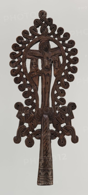 Processional cross, Ethiopian workshop. The pear-shaped frame depicts Christ on the cross with John and Mary standing at the foot of the cross in flat relief. The figures are schematically rendered. Christ is bearded, long-haired, with open eyes; his head is slightly tilted to his right left shoulder. He is dressed only in a loincloth and his feet are nailed with a single nail. The pear-shaped frame is decorated with a braided-rope pattern and bordered along the outer edge with an openwork braided ornament. The cone-shaped sleeve, flattened on the sides, is decorated with a vertical strip of braid and at the bottom with three horizontal strips of checkered, zigzag, and braided pattern. The arms connecting the sleeve with the cross take the form of an openwork braid. 20th century, Ethiopia. Cross; height 40.5 cm, width 16.0 cm., A carved wooden object in dark brown with a tall central figure flanked by two smaller figures, all carved in relief within an oval frame; the frame and surroun...