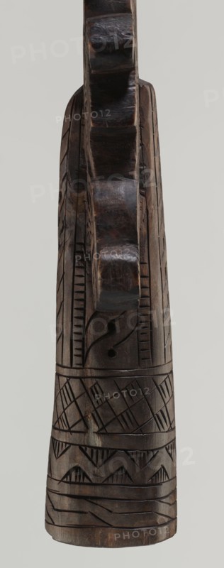 Processional cross, Ethiopian workshop. The pear-shaped frame depicts Christ on the cross with John and Mary standing at the foot of the cross in flat relief. The figures are schematically rendered. Christ is bearded, long-haired, and has open eyes; his head is slightly tilted to his right left shoulder. He is dressed only in a loincloth and his feet are nailed with a single nail. The pear-shaped frame is decorated with a pattern of braided rope and bordered along the outer edge with an openwork braided ornament. The cone-shaped sleeve, flattened on the sides, is decorated with a vertical strip of braid and at the bottom with three horizontal strips of checkered, zigzag, and braided pattern. The arms connecting the sleeve with the cross take the form of an openwork braid. 20th century, Ethiopia. Cross, height 40.5 cm, width 16.0 cm., A carved wooden object viewed from the front with a tall narrow form and a rounded top, showing a dark brown wood tone with lighter brown highlights and s...