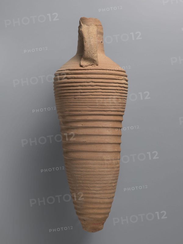 Amphora, unknown Egyptian workshop; amphorae; Egyptian ceramics; storage vessel; Coptic vessel;, amphora; Nile clay. Amphora with a spindle-shaped belly, fluted walls, and a pointed tip. Narrow neck with reflected rim, elongated and flowing into sloping shoulders. Two cylindrical vices attached to the neck and shoulders. A hole for wine fermentation drilled in the neck. Interior impregnated with resin. Outer surface light brown and smooth; ribbed belly. Made of light brown alluvial clay Naqlun AC1C ceramic fabric, 5th–7th century, Naqlun findspot. Vessel amphora, height 56.6+ cm preserved, neck diameter 6.5 cm, body diameter 20.2 cm., A tall, elongated ceramic vessel with a tapered base and a narrow top, showing a rough, textured surface with horizontal ridges running along its body; the vessel is primarily terracotta in color with variations of light brown and beige, and it rests against a smooth gray background.