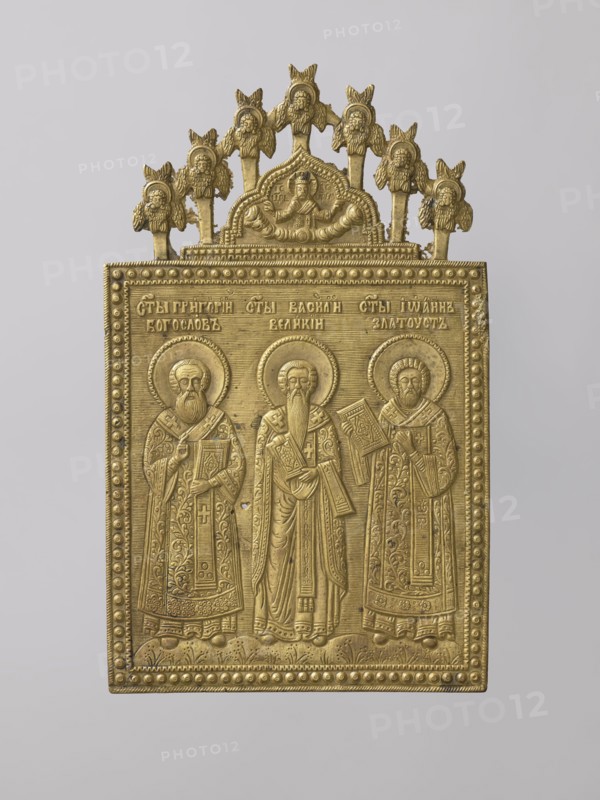 Icon: Holy Fathers of the Church. Old Believers workshop. Metal icon showing Saints Gregory the Theologian, Basil the Great, and John Chrysostom in full-figure, frontal views. They are dressed in bishops' robes, holding books, standing on flower-covered ground. Identifying inscriptions above them. Background horizontally engraved with thin lines. A ribbon ornament with raised dots surrounds the composition. On the icon's axis is a medallion topped with a kokoshnik-shaped arch depicting Christ as the High Priest, blessing with both hands. Cherubim and ripidions form a semicircular arch around it. Relief is precise and meticulous, with ornamentation and careful detail. 19th century, Russia. Materials: metal. Dimensions: height 19.8 cm, width 12 cm, depth 0.2 cm., A rectangular metal plaque with a decorative arched top, rendered in a gold color, showing three standing robed figures with halos, each holding a book or object, framed by a beaded border and a patterned lower edge; above the f...