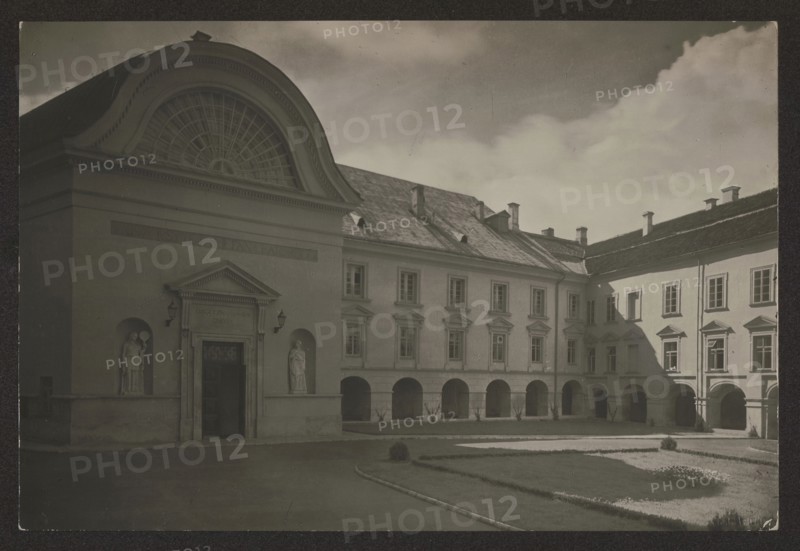 Vilnius. Stefan Batory University - Skarga Courtyard, Bulhak, Jan 1876-1950 photographer, Vilnius Lithuania, ca. 1934, photography, nature photography, height 11.3 cm, width 16.7 cm, A sepia-toned courtyard scene shows a large classical building with arched openings along the ground level and rectangular windows on the upper floor, a curved pediment with a fan-shaped window above a decorated entrance, and two niches flanking the doorway each containing a standing statue; a paved drive and formal lawn with trimmed hedges and planting beds occupy the foreground, and the building's sloped roof with small chimneys and dormers is visible under a sky with scattered clouds.