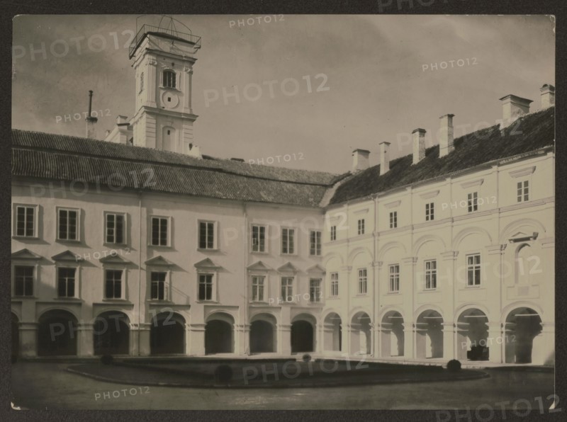 Vilnius. University - Skarga Courtyard, Bulhak, Jan 1876-1950 photographer, Vilnius Lithuania, ca. 1937, photography, nature photography, height 11.7 cm, width 16.2 cm, A courtyard framed by a large pale building with a tiled dark roof and a tall tower rising above it, showing a group of arched arcades at ground level and rows of rectangular windows above, with several chimneys projecting from the roofline; the courtyard has a low raised border around a central paved area and a few small rounded shrubs; visible colors include shades of white, cream, beige, dark brown, and gray.