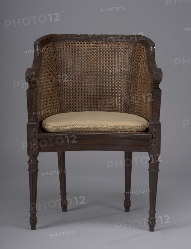 Armchair in the Louis XVI style; seat and backrest woven; 18th/19th century; height 81 cm, width 55 cm, depth 50 cm;, A wooden chair with a woven cane back and sides and carved wooden arms and legs, featuring a light beige cushion on the seat; visible colors include brown tones in the wood and cane, beige on the cushion, and a gray background.
