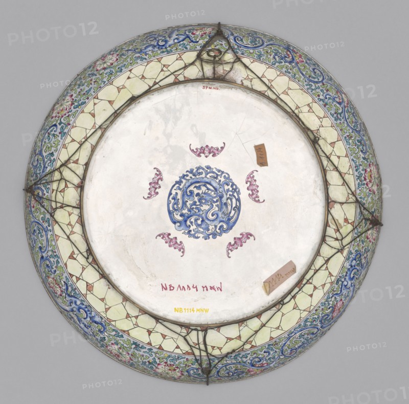 Platter, Large decorative platter with flat bottom and edges rising in an arc, set on a low, wide ring foot. Obverse: figural composition in a large tondo overlapping the lower parts of the slightly rising edges — a genre scene of scholars taking a siesta on a sunny summer day on a terrace of a landscape park. High foundations of decoratively bricked boulders rise above the valley of a large river. The terrace with an openwork balustrade is overgrown with grass, large deciduous trees, and banana trees nestled among rocks and boulders. In the center of the terrace is a large table with three scholars seated and a girl beside them. Reverse: mirrored leg decoration — a decorative composition of five pink bats surrounding a blue dragon, combined with plant motifs. 19th century, China. Plate; height 5.8 cm, diameter 44 cm., A round ceramic plate viewed from above with a central circular medallion decorated in blue surrounded by a group of ornamental pink motifs on a white background; the ri...