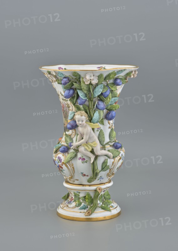 Vase, 19th/20th century, Meissen, vase, diameter 36 cm, diameter 24 cm base, The image shows a decorative porcelain vase with a flared rim and a rounded body on a pedestal, predominantly white with accents of gold, green, blue, purple, yellow, and pink; the vase is embellished with raised green leaves and clusters of blue-purple fruit, small white flowers, and painted floral motifs, and features a small sculpted cherub figure in pale flesh tones with yellow cloth elements clinging to the foliage; gold trim highlights the rim and base, and the background is a uniform grey.