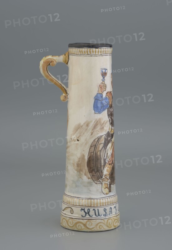 Mug. Cylindrical mug, slightly tapering toward the top, with a handle. Mantle decorated with an image of a nobleman sitting on a barrel holding a glass goblet in his right hand; a coat of arms next to the handle; plant decoration. Inscription on the pedestal: Hulaj dusza. Tankard; height 28 cm, width 15 cm with handle, inlet diameter 6.5 cm, bottom diameter 9 cm. Majolica manufactory in Nieborów manufacturer, 1881–1899. Maker: Jadwiga Hyzycka 1856–1922, 1881–1890., A tall ceramic tankard with a curved handle painted in shades of tan and ochre stands against a gray background; the body of the tankard is primarily off-white with painted figures in blue, brown, tan, and black, including a person raising a cup and parts of a wooden barrel, and decorative bands in yellow ochre and black near the top and base.