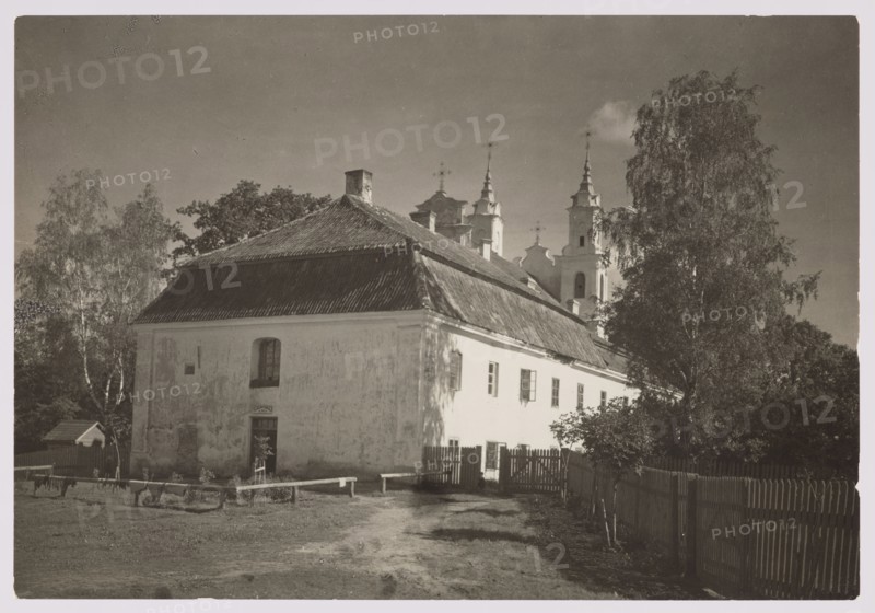 Calvary, Bulhak, Jan 1876–1950 photographer, Vilnius, ca. 1934. Calvary of Vilnius Werki; churches; architecture; sacred architecture; Vilnius Baroque style; photography; nature photography. Image size: height 11.5 cm, width 16.5 cm., A large light-colored two-story building with a steep dark roof fills the foreground, showing a worn exterior and several windows; a small door is visible on the nearer wall. A dirt path and grassy area lead up to the building, bordered by a wooden fence and a few low benches or rails. Tall trees with leafy canopies stand to the left and right of the building, casting shadows. Behind the building rise ornate towers with pointed spires partly obscured by the roof and trees. The scene appears in muted tones of black, white, gray, and sepia.