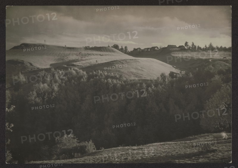 Ponarskie Mountains above the Wilja River Gorzuny, Jan Bulhak 1876–1950, photographer. Vilnius environment, Vilnius Lithuania. Nature photography; landscapes, hills. ca. 1934. Photography. Height 11.6 cm, width 16.6 cm. Purchase provenance., A landscape showing rolling hills and a wooded valley under a cloudy sky, with a group of houses along a ridgeline; visible colors include shades of black, gray, white, and muted sepia tones.
