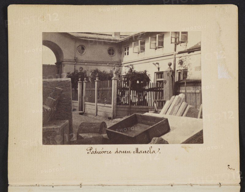 The Courtyard of the Mancel House View of the courtyard of the house and stonemason-sculptor workshop of Jan Józef Manzel, photographed by Karol Beyer 1818–1877; depicts Jan Józef Manzel 1806–1875. Date: 1861–1863. Medium: photographic print, nature photography/photography. Image: 5.5 cm high x 8 cm wide; support: 11.3 cm high x 8.8 cm wide. Location depicted: Warsaw, Chlodna Street, Masovian Voivodeship., A sepia-toned photograph shows a courtyard scene framed by a light-colored mat with a darker border. In the image, a building facade with a covered archway and round windows sits in the background, and a row of open rectangular windows is visible along the upper level. A wooden picket fence with decorative posts and finials runs across the middle ground, partially obscured by foliage and small trees. In the foreground there are several stone blocks and slabs, including a rectangular wooden or stone trough tilted on its side and a few angled slabs leaning against each other. Visible c...
