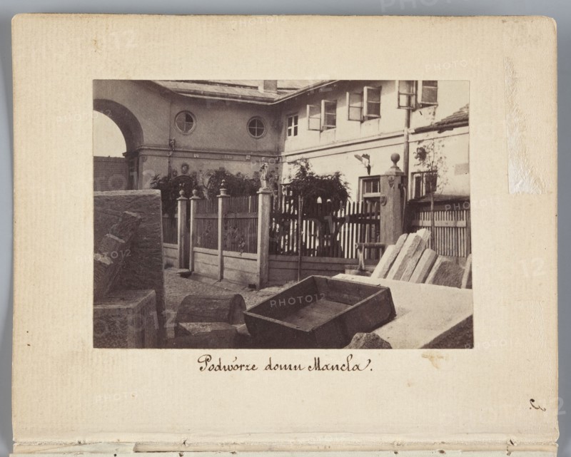 The Courtyard of the Mancel House View of the courtyard of the house and stonemason-sculptor workshop of Jan Józef Manzel, photograph by Karol Beyer 1818–1877; depicts Jan Józef Manzel 1806–1875 and his workshop; dates: 1861–1863; location: Chlodna, Warsaw Masovian Voivodeship; medium: photographic print, nature photography/photography; print dimensions: 5.5 cm x 8 cm; support dimensions: 11.3 cm x 8.8 cm., A sepia-toned photograph mounted on a beige card shows an outdoor scene with a courtyard enclosed by a wooden fence and gate. In the foreground there are several wooden and stone objects including a rectangular wooden box, a few planks or stone slabs leaning at an angle, and a rounded wooden barrel-like piece. Behind these objects a picket-style fence with vertical slats and decorative posts separates the courtyard from a building. The building has a pale facade with multiple windows, some open, and circular windows on an upper section. There is an archway on the left side of the bu...