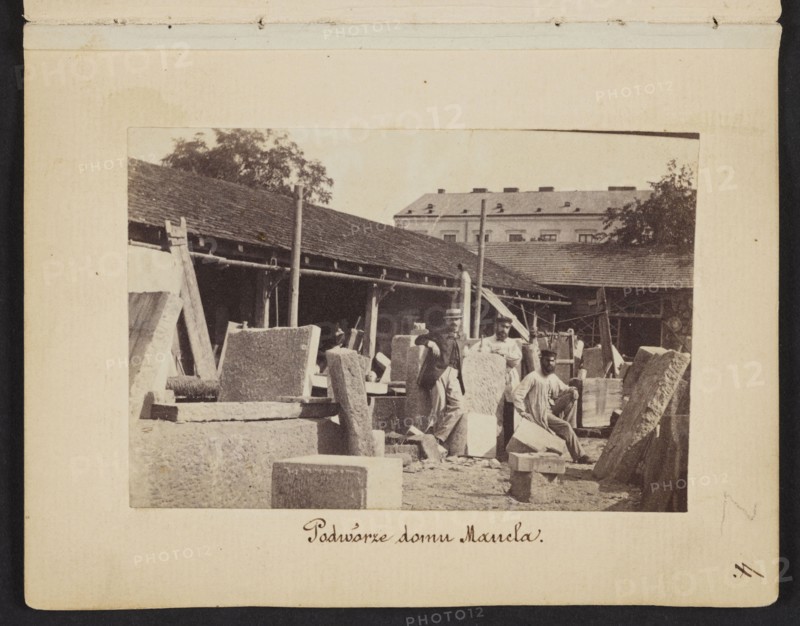 The Courtyard of Mancel's House View of the stonemasonry and sculpture workshop of Jan Józef Manzel. In the center the sculptor Józef Manzel, the owner's son, photograph by Karol Beyer 1818–1877; subjects: Jan Józef Manzel 1806–1875, Józef Manzel 1834–1904; other photographer: Leopold Méyet 1850–1912. Date: 1861–1863. Place: Warsaw Chlodna street. Genre: photography, nature photography; depiction: sculptors, sculptors' workshop, iconography. Photographic print: 5.7 × 8.4 cm; support: 11.3 × 8.8 cm., The image shows a sepia-toned scene of an outdoor yard with a group of several people among many large stone blocks and carved slabs, a few leaning against one another and several scattered on the ground; a long single-story building with a sloped roof runs along the background, with a taller multi-story building partially visible behind it and some trees rising above the roofs; a few vertical poles and wooden supports stand among the stones, and one person sits on a block while others stan...