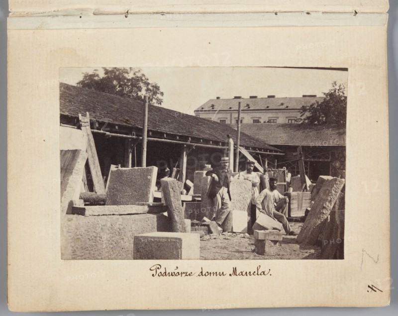 The Courtyard of Mancel's House View of the stonemasonry and sculpture workshop of Jan Józef Manzel. In the center the sculptor Józef Manzel, the owner's son, photograph by Karol Beyer 1818–1877, depicting sculptors Jan Józef Manzel 1806–1875 and Józef Manzel 1834–1904; 1861–1863; place: Warsaw, Chlodna Street; genre: photography, nature photography; photographic print: 5.7 × 8.4 cm; support: 11.3 × 8.8 cm., The image shows an outdoor scene with a group of people among large stone blocks and slabs in a yard bordered by a long, low building with a pitched roof and exposed beams. A few men are standing or sitting on the stones; one man wears a hat and a coat, others wear lighter shirts and work clothes. There are several upright and leaning stone pieces, wooden supports, and simple scaffolding or poles around the work area. In the background a larger multi-storey building and some trees are visible. The visible colors are sepia tones, beige, brown, and muted gray.