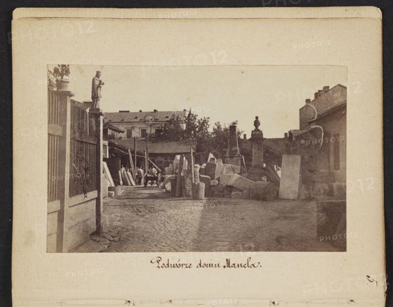 Courtyard of Mancel's House View of Jan Józef Manzel's stonemasonry and sculpting workshop, photograph by Karol Beyer 1818–1877; depicts Jan Józef Manzel 1806–1875, Léopold Méyet 1850–1912; subject: sculptors' workshop, tombstones; place: Chlodna, Warsaw, Masovian Voivodeship; date: 1861–1863; medium: photographic print nature photography; dimensions: print 5.7 × 9.2 cm; backing 11.3 × 8.8 cm., The image shows an outdoor courtyard scene with a cobblestone ground and a pathway leading between structures; there is a tall fence on the left with a statue mounted on a post and a group of rectangular stone blocks and slabs stacked and scattered toward the center and right, accompanied by a few upright pillars and carved stone elements; a few people are visible near the middle distance beside a cart and wooden beams, and there are buildings with pitched roofs and trees in the background; the visible colors include shades of brown, beige, tan, sepia, and muted gray.