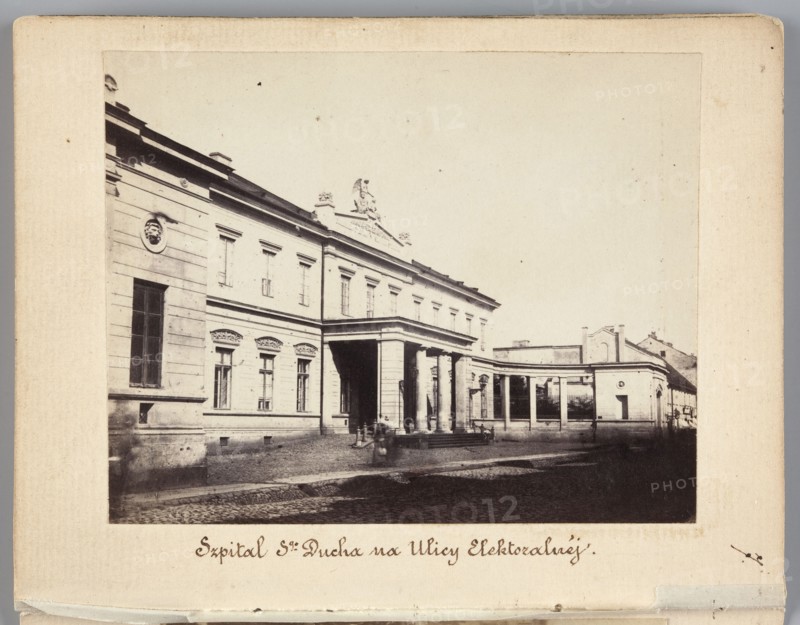 Hospitium Sancti Spiritus Hospital of the Holy Spirit on Elektoralna Street, Warsaw. Building designed by architect Józef Orlowski 1819–1880. Photograph by Karol Beyer 1818–1877, c. 1861–1863. Subject: hospital, Elektoralna Street, Warsaw St. Ducha. Medium: photographic print; image 7.0 × 8.7 cm; support 11.3 × 8.8 cm. Nature photography., A sepia-toned photograph shows a classical building with a facade featuring tall windows, decorative stonework, and a portico supported by columns; a group of columns extends along a curved colonnade to a lower wing on the right. The building surface appears in shades of beige and brown, with darker brown shadows in recessed areas and the foreground cobblestone street. A few indistinct figures stand near the portico. The sky is a pale off-white, and the photograph is mounted on a light cream mat with a slightly darker border.