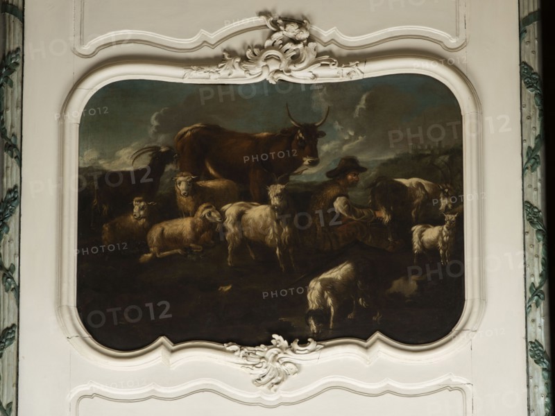 Shepherd's Scene with a Shepherd Milking a Goat. The overall tones are browns and dark blues. The surface is fully composed, the focal point is a cow standing at the back and a group of five animals—three sheep and two goats—filling the right side of the scene. On the right, a seated shepherd in a sheepskin coat and hat milks a goat into a wooden bucket. Next to the goat is a small white kid. At the front a dog laps water from a shallow pond. In the background on the right, city buildings are visible on a hill beneath a dark, puffy-clouded sky. In the upper corner is the Temple of the Sibyl in Tivoli. Several sheep and rams share identical faces. Philipp Peter Roos, known as Rosa di Tivoli born August 30, 1657 in Sankt Goar; died January 17, 1706 in Rome — German landscape painter; the painting may be by him possibly related to NB 4918, NB 4920, and NB 4921. 18th century, Italy; overdoor; oil on canvas; height 91.0 cm, width 127.0 cm., A painted scene set within an ornate white frame s...