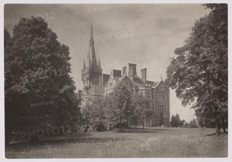 Lentvaris palace; photographer Jan Bulhak 1876–1950; architecture; Lentvaris, Lithuania; palaces; brick architecture; ca. 1934; nature photography/photography; height 11.7 cm, width 16.8 cm; purchase provenance., A large stone building with multiple steep roofs and a tall pointed central spire sits partly obscured by a group of trees in the foreground; several chimneys and decorative roof details are visible on the building. The scene shows a grassy lawn with scattered small flowers in front, bordered by a few large mature trees on both the left and right sides. The sky above is light with subtle cloud texture. Visible colors include shades of gray, black, and white.