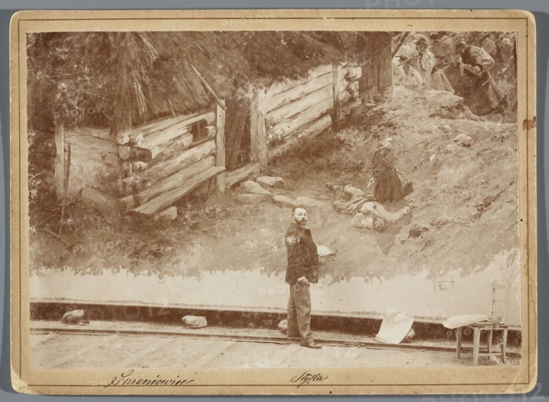 Painting the Raclawice Panorama: Jan Styka standing with a palette next to a part of the canvas depicting a woman mourning a killed peasant, in front of a cottage in Smoniowice at the Dziemierzyce crossroads. Hamel i Feigl Lviv; typesetting of photographic materials; 1892–ca.1905 publisher. Reproduction of painting; Styka, Jan 1858–1925 and others; Kossak, Wojciech 1856–1942 reproduction. Raclawice Panorama, Polish painting; painters' studios; portraits of artists; painting panoramas; painting accessories; painting, 1893/1894; Lviv. Photography from nature; photographic print: height 11.8 cm, width 17.2 cm; print glued onto cardboard backing: backing height 13.5 cm, width 18.5 cm., A sepia-toned scene shows a large painted backdrop of a rustic outdoor setting with rough wooden logs forming a structure and a thatched roof, textured ground with rocks and vegetation, and a few women in period clothing on a slope, one seated and another standing nearby; in front of the painting stands a ma...
