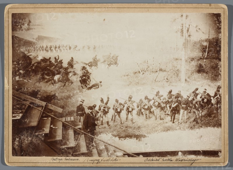Painting the Raclawice Panorama: Teodor Axentowicz standing on scaffolding by a section of the canvas depicting a battle scene of the company of Józef Wodzicki’s 2nd Infantry Regiment; photographic print photography from nature, glued to cardboard backing; 1893/1894; image height 11.9 cm, width 17.3 cm pad 13.5 × 18.5 cm. Depicts painters and painting accessories, scaffolding, and artists' studios; related to the Panorama of the Battle of Raclawice by Wojciech Kossak and Jan Styka; Teodor Axentowicz 1859–1938, Wojciech Kossak 1856–1942, Jan Styka 1858–1925., The image shows a sepia-toned scene with a group of soldiers in uniform standing and marching on grassy terrain, a group of mounted cavalry in motion with horses and riders to the left, and a few figures in civilian clothing near wooden structures or steps in the lower left. The background includes trees and open ground, and the overall colors visible are shades of brown, tan, beige, and off-white.