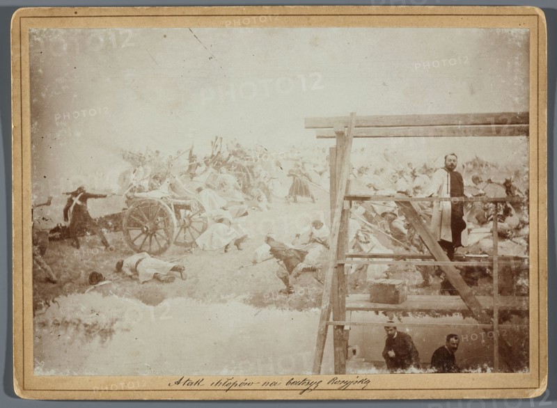 Painting the Raclawice Panorama: Jan Styka standing on scaffolding by a section of the canvas depicting the capture of a battery of Russian cannons by the leading group of the first column of scythe-bearers, Hamel and Feigl; photographic print glued to cardboard; ca. 1893–1894; photographer, reproduction of Wojciech Kossak and Jan Styka; subjects include panorama painting, artists' studio, scaffolding; dimensions of photographic print 11.9 × 17.1 cm, cardboard 13.5 × 18.5 cm., The image shows a pale, sepia-toned scene with a large painted or staged battle on the left and a wooden scaffold structure on the right; the battle area contains a group of figures in light-colored clothing near a wheeled cart, with some figures lying on the ground and others in dynamic poses, and darker-clothed figures appearing among them; the scaffold has a few men standing or crouching on its platforms, one man in a light coat standing near the upper railing, and two men lower down; visible colors include be...