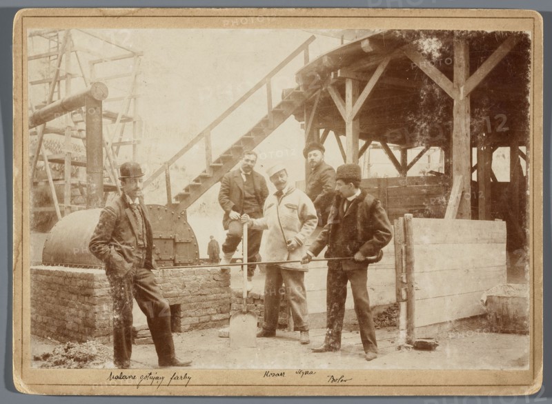 Painting the Raclawice Panorama: painters mixing paints in the rotunda; from the left: Zygmunt Rozwadowski, Ludwig Boller, Wojciech Kossak, Jan Styka, Tadeusz Popiel, Hamel and Feigl, photographic print, ca. 1893–1894; print glued to cardboard backing. Photography of artists in a studio depicting painting panoramas, scaffolding, painting accessories; dimensions of photographic print 11.8 × 17.4 cm, cardboard 13.5 × 18.5 cm., A sepia-toned photograph shows a group of men standing and posing in an outdoor industrial or construction setting with wooden beams and a raised wooden platform with stairs in the background; a few men wear hats including a bowler and a fur hat, and their clothing includes coats and suits in muted tones; one man leans against a low brick structure near a cylindrical metal drum, another man holds a shovel, and another holds a long metal rod across a low wooden barrier; there are wooden posts, planks, and scaffolding visible, with dirt or gravel on the ground, and t...