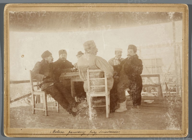 Six painters seated at a table on the central platform in the rotunda painting the Raclawice Panorama; from the left: Tadeusz Popiel, Jan Styka, Wlodzimierz Tetmajer, Michal Sozanski back, Wojciech Kossak, Ludwig Boller, Hamel and Feigl photographic studio, ca. 1893–1894. Photography, print glued to cardboard, photographic print: 11.7 × 17.3 cm; cardboard: 13.5 × 18.5 cm. Subjects and artists: Panorama of the Battle of Raclawice; painters’ studio, scaffolding, portraits of artists. Identified persons: Kossak, Wojciech 1856–1942; Styka, Jan 1858–1925; Popiel, Tadeusz 1863–1913; Sozanski, Michal 1853–1923; Boller, Ludwig 1862–1896; Tetmajer, Wlodzimierz 1861–1923., A sepia-toned photograph showing a group of men seated around a small table under a covered wooden structure, the scene dominated by brown, beige, cream, and muted gray tones; the men wear a mix of dark and light outer garments and hats in shades of black, brown, and gray, and one man facing away wears a lighter-colored coat w...