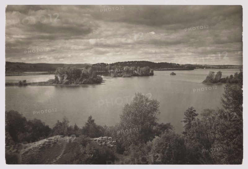 Trakai - islands on the lake; Bulhak, Jan 1876-1950 photographer; Trakai Lithuania; lakes; landscapes; ca. 1934; nature photography; height 11.5 cm; width 17.0 cm, A sepia-toned photograph showing a wide body of water with a few small wooded islands and a shoreline lined with trees, a group of taller trees in the foreground partially obscuring the water, gently rippled water surface, distant low hills on the far shore, and a sky filled with layered clouds in various shades of brown and beige.