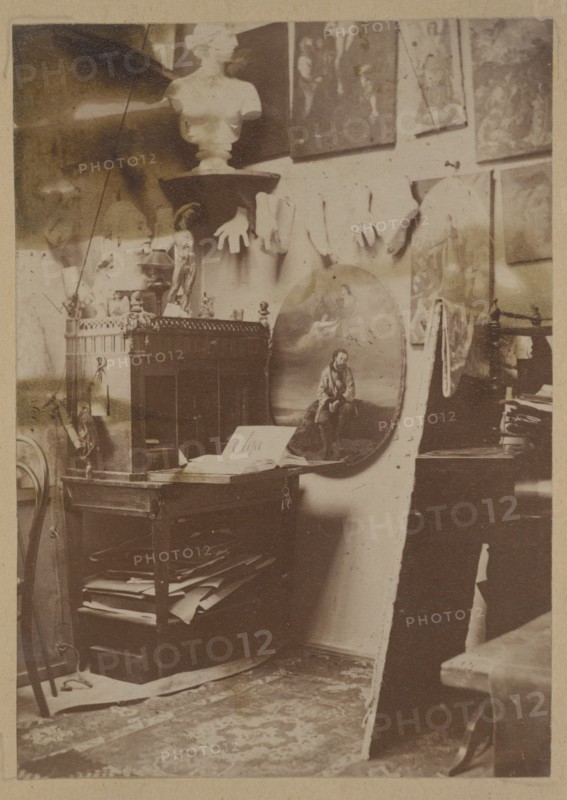 Ignacy Jasinski's 1833–1878 painting studio at the Hotel Europejski in Warsaw; Ignacy Jasinski 1833–1878, photograph, print glued across the entire surface on the verso page of the 22nd page of an album, 1869–1878; subject: interiors, artists' studio, painters; medium: photography nature photography/photograph in an album; dimensions: height 10.3 cm, width 7.3 cm; Polish painting reproduction/iconography., A cluttered interior scene with a wooden table or cabinet holding books and papers, a round painted panel leaning against the wall depicting a seated figure, a bust on a pedestal near the top, several gloves hanging on the wall, framed artworks arranged on the wall, a few sculptural objects and small statuettes on the furniture, a patterned rug on the floor, and visible tones of brown, beige, sepia, cream, and hints of dark gray.