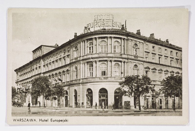 Hotel Europejski at Krakowskie Przedmiescie, Warsaw; photographer, published by Wydawnictwo Salonu Malarzy Polskich Kraków; architecture; hotels; Hotel Europejski Warsaw; Warsaw Masovian Voivodeship; Polish architecture; neon signs; ; postcard glued to the backing with two strips on the shorter side; 1939/1940; Krakow; postcard; height 9 cm; width 14 cm, A black-and-white photograph of a large multi-story corner building with classical architectural features, including arched ground-floor openings, tall rectangular and arched windows, pilasters, and decorative cornices; a rooftop metal sign structure is visible atop the curved corner section. The facade shows a series of repeated window bays extending along both street-facing sides, with a few trees lining the sidewalk in front of the building and a group of people walking or standing near the entrances. The street in front appears wet and reflects light, and the overall scene includes tones of black, white, and various shades of gray.
