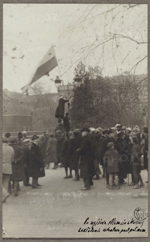 Planting a white-and-red flag in front of the Governor's Palace by a student of the University of Warsaw, after the Germans left the city; photographer: Waclaw Saryusz-Wolski 1870–1933; World War 1914–1918; Warsaw, Governor's Palace, Krakowskie Przedmiescie; Year: 1918; genre scene; national flags Poland; University of Warsaw founded 1816; photograph on postcard paper glued with upper edge to cardboard; board: Polish Legions 1914–1918, part II, 10/11/1918; photographic print: height 13.9 cm, width 8.6 cm; base: height 24.6 cm, width 16.7 cm., A group of people gathered outdoors on a street or square, most wearing dark coats and hats, with a few lighter-colored coats visible; several adults and some children are standing together, some facing different directions. In the background there are leafless tree branches extending across the upper part of the scene and a dense group of trees or shrubs behind the crowd. A tall lamp post is visible with a person standing or climbing on it, and a...