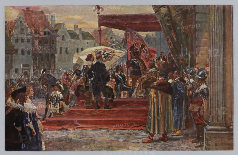 Wojciech Kossak 1856–1942, The Predatory Fief: The Prussian Homage, 1912; page from the Prussian Spirit portfolio, reproduction of a work of art after a painting; print 17.7 × 27.6 cm mounted on cardboard 29.0 × 39.7 cm; album/portfolio of reproductions; historical scene; Polish painting; publisher: Zaklady Graficzne B. Wierzbicki i S-ka Warsaw., A crowded outdoor scene shows a raised red platform with a canopy where a group of figures in historical dress are gathered. Several people wear dark blue, black, brown, and gold garments, including cloaks, capes, and fur-trimmed coats, while a few wear metallic armor with silver helmets. There are banners and large cloths in white and pink tones behind the central figures, and a seated person in darker clothing appears near the center of the platform. In front of the platform a red carpet or cloth covers steps leading down to a cobbled ground that is gray and brown. To the left, a group of onlookers in dresses and hats in shades of blue, whit...