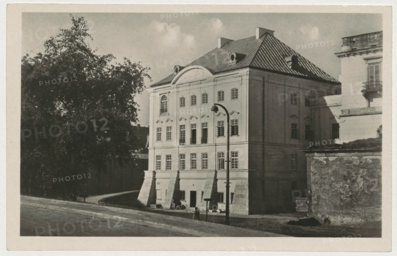 Warsaw. Socialist construction resurrects and discovers the beauty of neglected works of architecture, 1948, photographer Jan Bulhak 1876–1950; postcard; photographic postcard; Palac pod Blacha Palace under the Tin Roof, Warsaw Masovian Voivodeship; height 14 cm, width 9 cm., A street view showing a large multi-story building with a pitched roof and dormer windows next to a smaller section of another building, a tall tree with dense foliage partially obscuring the left side, a curved pathway or road in front, a tall lamppost near the building, a low textured wall or rubble area to the right, a few small figures near the building entrance, and clouds in the sky; visible colors include shades of black, white, gray, and sepia tones.