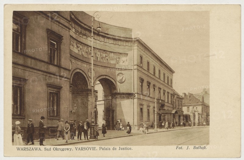Warsaw. District Court. Varsovie. Palais de Justice, Bulhak, Jan 1876-1950 photographer, Tanski, Jerzy, Pac Palace Warsaw, Warsaw Masovian Voivodeship, ca. 1925, Warsaw, postcard, height 9 cm, width 14 cm, A sepia-toned street scene showing a large classical building with an arched entrance and decorative relief along the curved upper facade, flanked by rows of tall windows; a tall ornate lamppost stands near the arches. Several people in period clothing are gathered and walking along the sidewalk in front of the building, including men in hats and women in long dresses. The street extends to the right with additional buildings and shopfronts receding into the distance. Visible colors include shades of brown, beige, tan, and cream.