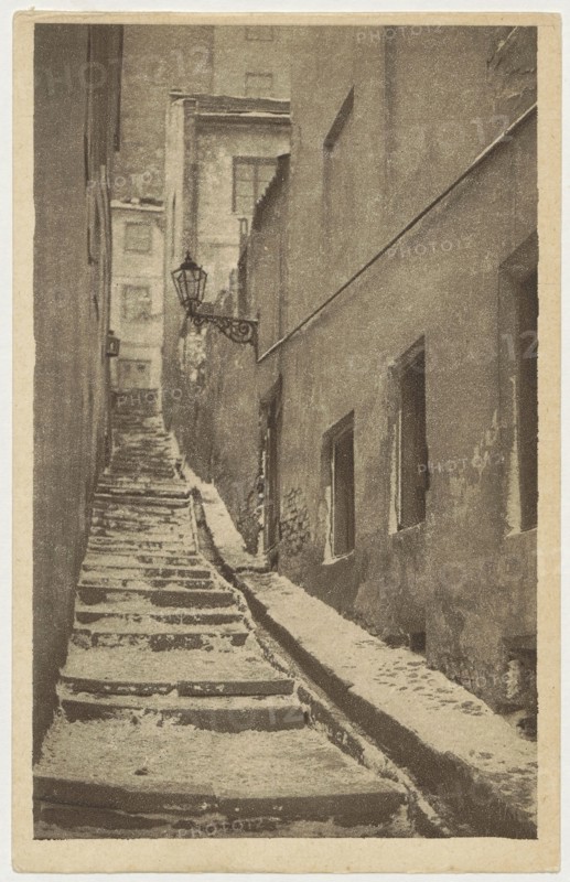 Warsaw. Old Town - Stone Stairs, St. Adalbert's Printing and Bookstore; Polish Society of Railway Bookstores Ruch publisher; Bulhak, Jan 1876–1950, photographer; ca. 1930; Stone Stairs, Warsaw street, Warsaw Old Town; postcard; 14 cm × 9 cm., A narrow, steep alleyway with worn stone steps leading upward between tall buildings, showing a rough stone path and a sloped gutter alongside the steps; the right building has several recessed windows and a textured, weathered wall, while the left side shows vertical building faces and a distant facade at the top of the alley; a decorative metal lantern extends from the left side near the middle of the scene. The image appears in muted tones of brown, beige, and sepia.