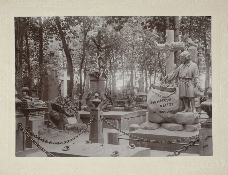 Tombstone of the composer Ignacy Komorowski 1824–1857 at Powazki Cemetery, Warsaw. Photograph glued across the entire surface onto a backing, ca. 1900. Nature photography/photograph, height 17.7 cm, width 24.8 cm. /photographer., The image shows a cemetery scene with a group of stone monuments and statues among trees; a stone sculpture of a young figure stands beside a large carved rock, and a tall cross with draped cloth is visible nearby; several upright tombstones and decorative urns occupy the area, with a low stone wall and chained posts enclosing part of the foreground; the ground is uneven with scattered stones and carved reliefs, and tree trunks and leafy branches form a wooded backdrop. The visible colors are shades of gray, sepia tones, off-white, and muted brown.