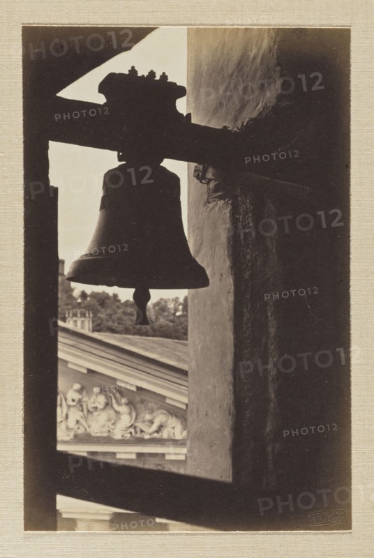 Vilnius. Cathedral bell tower; photograph in the album Vilnius Temples, volume 1, Jan Bulhak 1876–1950, photographer; pictorialism; cathedral Vilnius; photography of bells; 1912; Vilnius; photograph, photographic print: 15.3 × 10.0 cm and 15.5 × 10.0 cm; backing: 31 × 22.5 cm; album page: 31.1 × 23.5 cm, A sepia-toned photograph showing the dark silhouette of a bell hanging from a horizontal beam inside a window-like opening, with the bell's clapper visible and the bell's outline contrasted against a lighter background; to the right a textured vertical wall in shadow runs through the frame, and beyond the opening a portion of an ornate building facade with sculpted relief and a roofline is visible along with a group of trees, all depicted in shades of brown, beige, and dark sepia.
