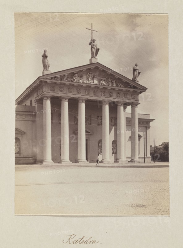 Cathedral; photograph, Bulhak, Jan 1876–1950, Vilnius Lithuania, cathedral Vilnius, 1912, photograph, photographic print: height 14.8 cm, width 11.2 cm, album page: height 31.0 cm, width 23.5 cm, A large classical building with a portico supported by a group of tall columns stands centered in the image, topped by a triangular pediment with sculptural relief and three statues along the roofline, one holding a cross; the façade and columns appear in shades of off-white and light beige, with shadows creating darker beige and gray tones. The sky behind the building shows muted gray and pale beige clouds. In front of the building is a wide open paved area in pale beige, and a small dark figure is visible near the base of the columns. Along the left and right edges of the building are architectural extensions in similar off-white and beige tones, and a few darker vertical elements and foliage appear at the far right in muted green-gray. The overall image has a sepia-like tint with warm beige...