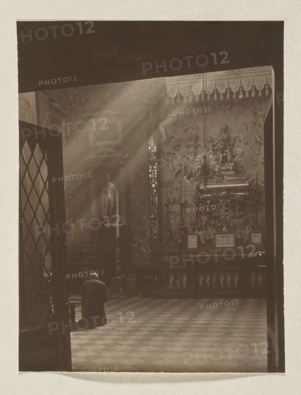 Cathedral. St. Casimir's Chapel; photograph, 1914. Photographer: Jan Bulhak 1876–1950. Subject: St. Casimir's Chapel in the Vilnius Cathedral Baroque architecture. Medium and format: photographic print, height 15.0 cm, width 11.4 cm; mounted on album page, height 31.0 cm, width 23.5 cm., A sepia-toned interior scene shows a person kneeling on a patterned floor in front of an ornate altar area. Sunlight streams through high windows creating visible beams that illuminate dust in the air. The altar wall features sculpted reliefs, a chandelier, decorative columns, and a raised platform with framed objects. A tall candle stand and a statue in a niche are visible near the altar. A partially open doorway with a diamond-patterned glass panel frames the left side of the scene. Colors visible are brown, beige, and muted gold tones.