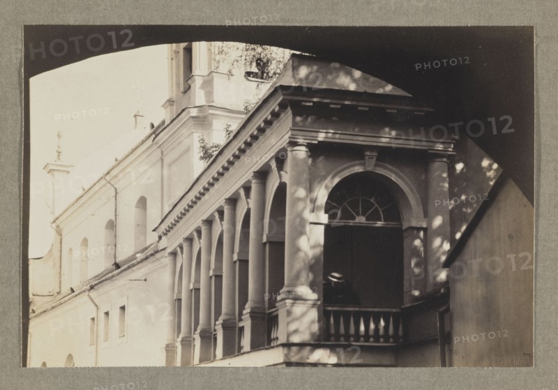 The Gate of Dawn; photograph, 1912, Vilnius; photographer Jan Bulhak 1876–1950; photographic print: 11.0 × 16.5 cm; album page: 31.0 × 23.5 cm., The image shows an exterior view of a building with a colonnaded balcony and arched openings; the facade displays columns, balustrades, and an arched window with a fanlight set above a dark opening where a person wearing a hat is partially visible; light and shadow create dappled patterns on the walls and columns; colors visible include shades of beige, cream, gray, and dark brown/black.