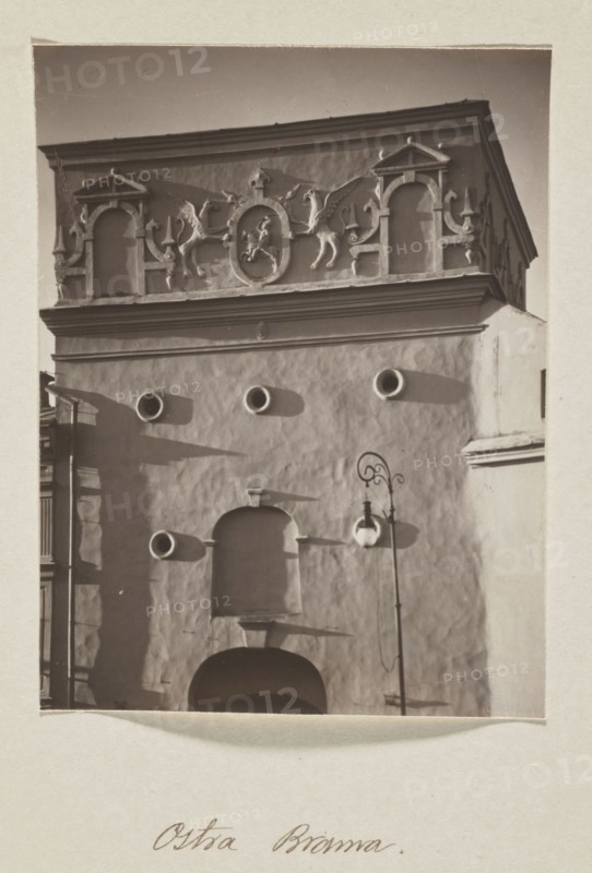 The Gate of Dawn; photograph, 1912. Photograph in the album Vilnius, volume 1, Jan Bulhak 1876–1950. Vilnius. Photographic print: 13.6 × 10.5 cm; album page: 31.0 × 23.5 cm., A vertical sepia-toned photograph shows the façade of an ornate building with a textured light-colored wall and a decorative upper frieze featuring sculpted figures and architectural niches. The top section has sculptural reliefs including winged creatures and framed medallion shapes, with ornamental columns and arch-like elements in pale tones. Below, the plain wall contains a group of circular openings that cast darker circular shadows, and a recessed arched niche above a larger dark arched doorway. A tall street lamp with a curved ornate arm and a globe-shaped lantern stands in front of the wall, casting a shadow. The image border is pale and the overall palette consists of brown, beige, cream, and darker brown shadows.