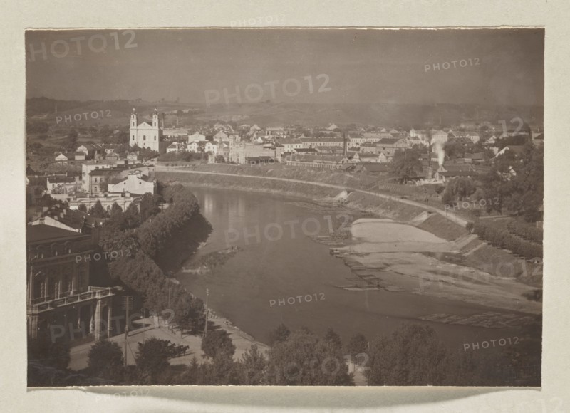 Wilia towards the Green Bridge; photograph, Wilno album, volume 1; Jan Bulhak 1876–1950; Vilnius panoramas; Wilia river, panorama of Vilnius; 1912; photograph, photographic print: 11.0 × 16.0 cm; album page: 31.0 × 23.5 cm, The image shows a riverside urban scene with a broad river curving through the center, bordered by tree-lined embankments and sandy exposed banks. Along the near bank there is a promenade area with a few trees and a large building with arches and a balcony. Across the river a group of buildings rises on gently rolling land, including a prominent twin-towered church with a light facade and several clustered structures of varied shapes and rooflines. There are a few smaller houses, some industrial-looking buildings emitting a plume of smoke, and a mix of open spaces and rows of trees. The foreground and riverbanks feature darker greens and browns, the buildings display muted light tones, and the sky appears a soft grayish tone.
